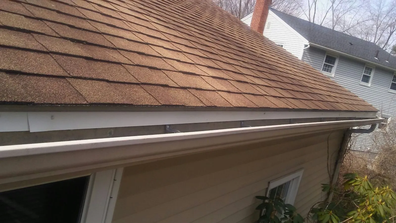 White seamless gutter installed along the shingled roofline of a tan-sided home