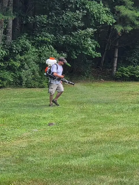 Technician crossing a broad lawn at the tree line with a backpack mosquito sprayer